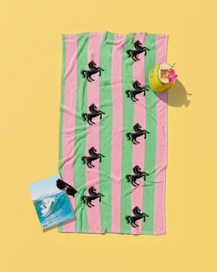 A pink and green striped beach towel printed with black horses, on yellow sand