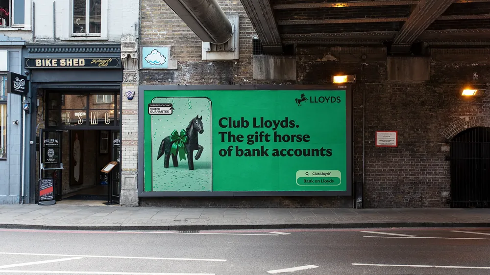 Club Lloyds billboard under a railway bridge: the gift horse beside the line “Club Lloyds. The gift horse of bank accounts”